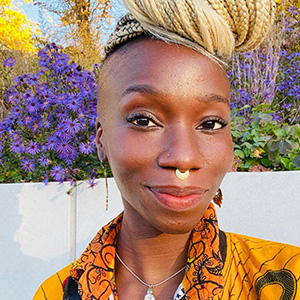 A smiling woman with braided hair piled in a topknot posing in front of purple flowers outdoors.