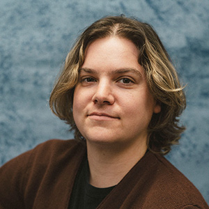 Headshot of Sunny Zimmerman, a person with shoulder-length wavy hair against a blue background.