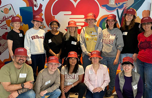 A group of volunteers wearing matching red "Vassar Serves" bucket hats posing together in front of a colorful wall.