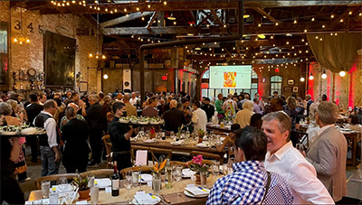 A crowded indoor event in a rustic hall with string lights, exposed brick walls, and long dining tables.