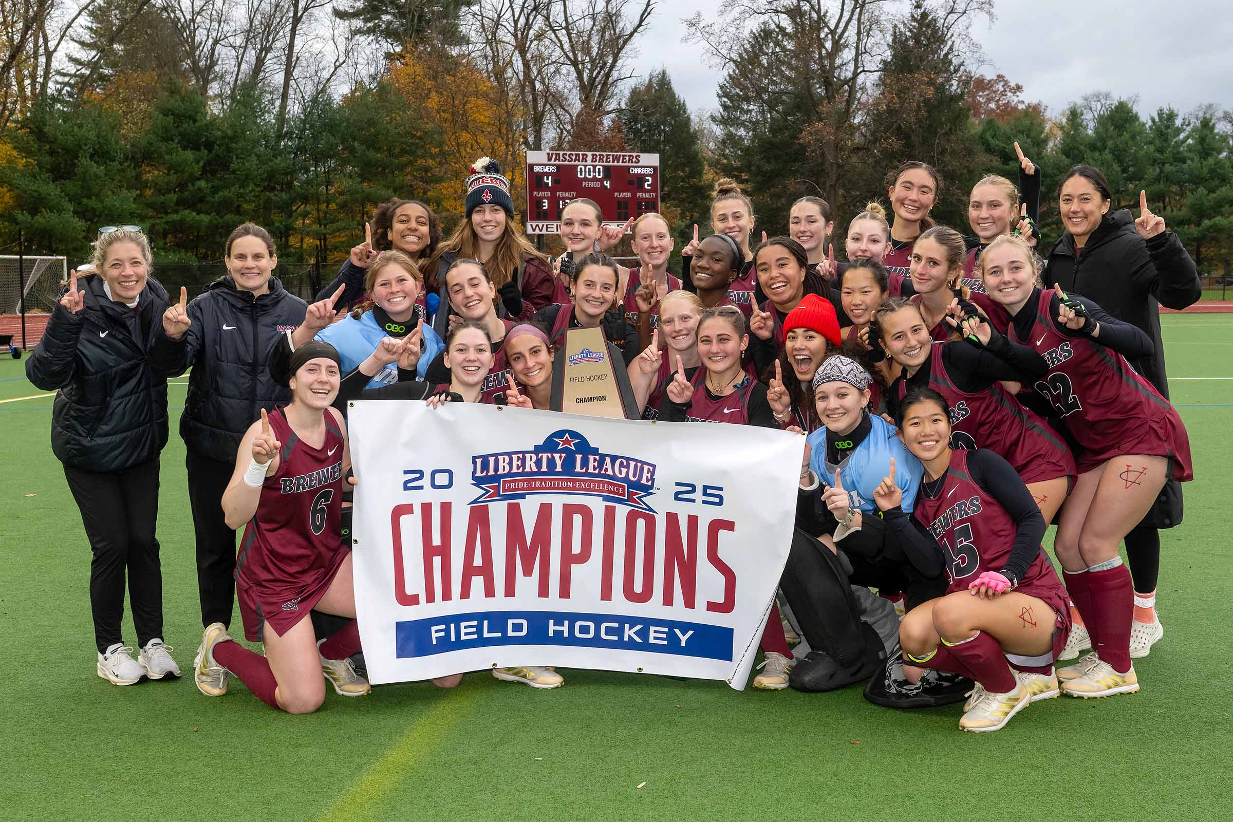 The Vassar women's field hockey team poses in their maroon uniforms on a turf field, holding a trophy and a banner that reads '2025 Liberty League Champions Field Hockey.'