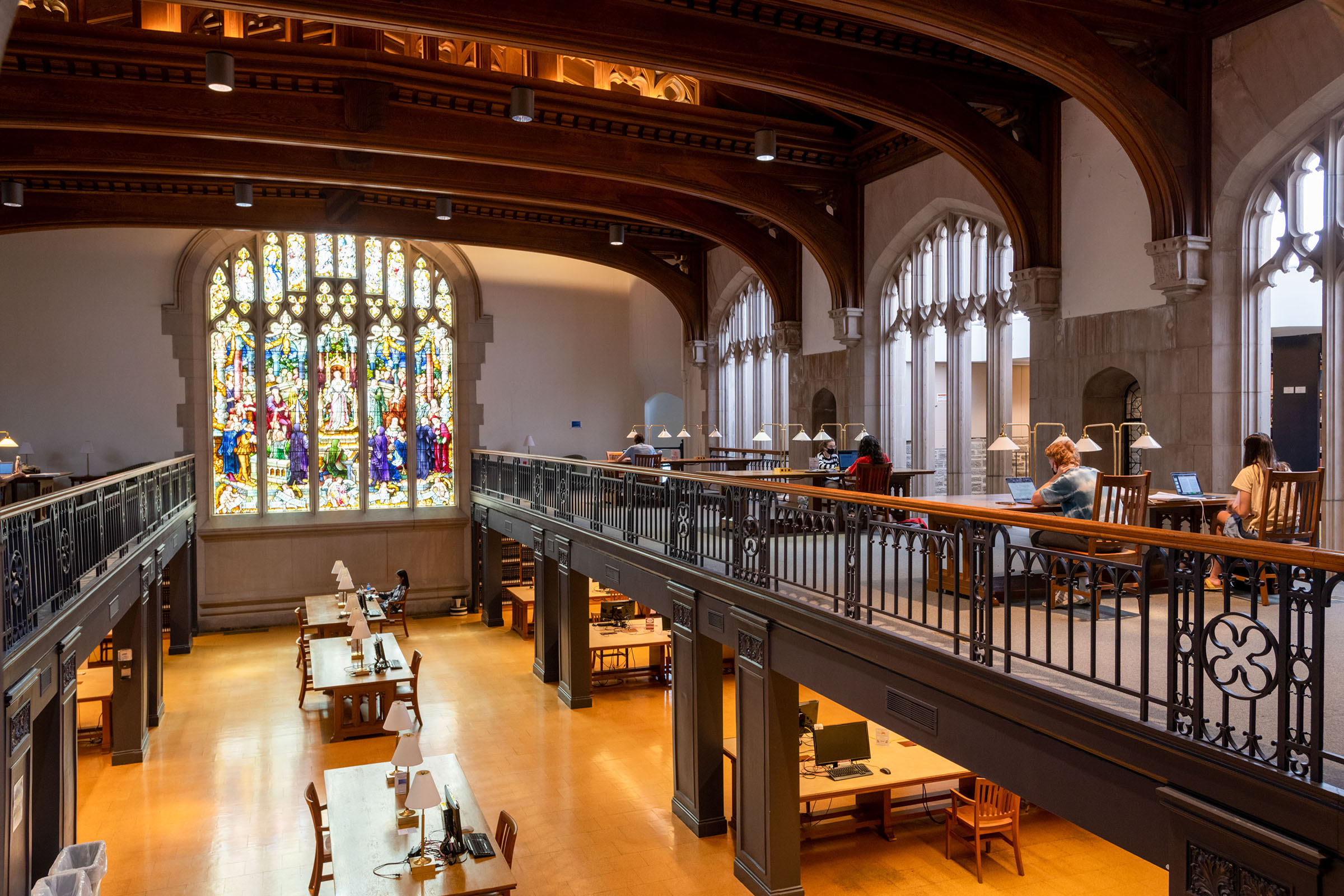 The a two-story interior view of a gothic-style library, with tables on the second floor overlooking the first floor, and a two-story stained glass window in the rear of the room.