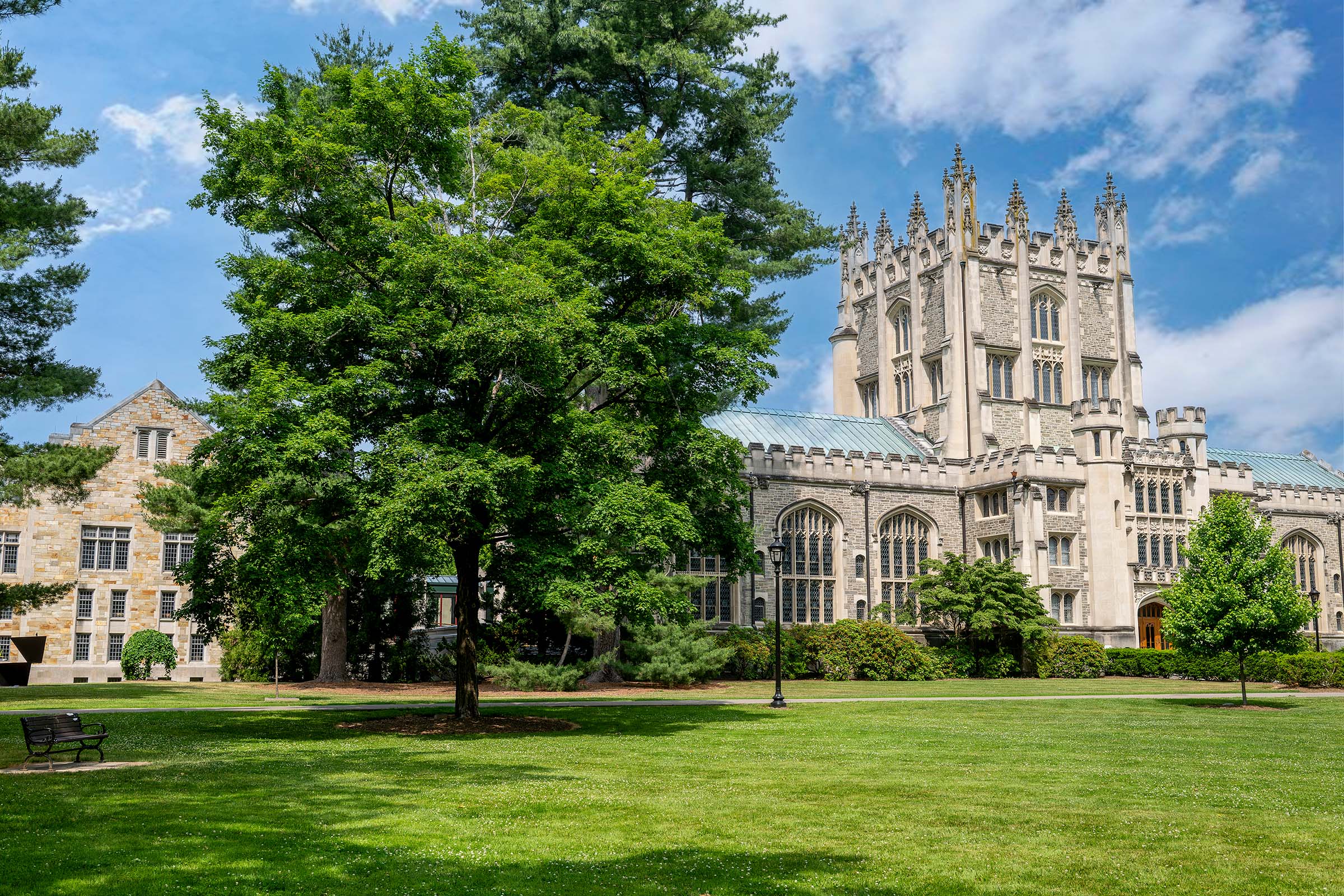 A Gothic-style stone building with a prominent ornate tower sits behind a lush green lawn with mature trees under a blue sky.