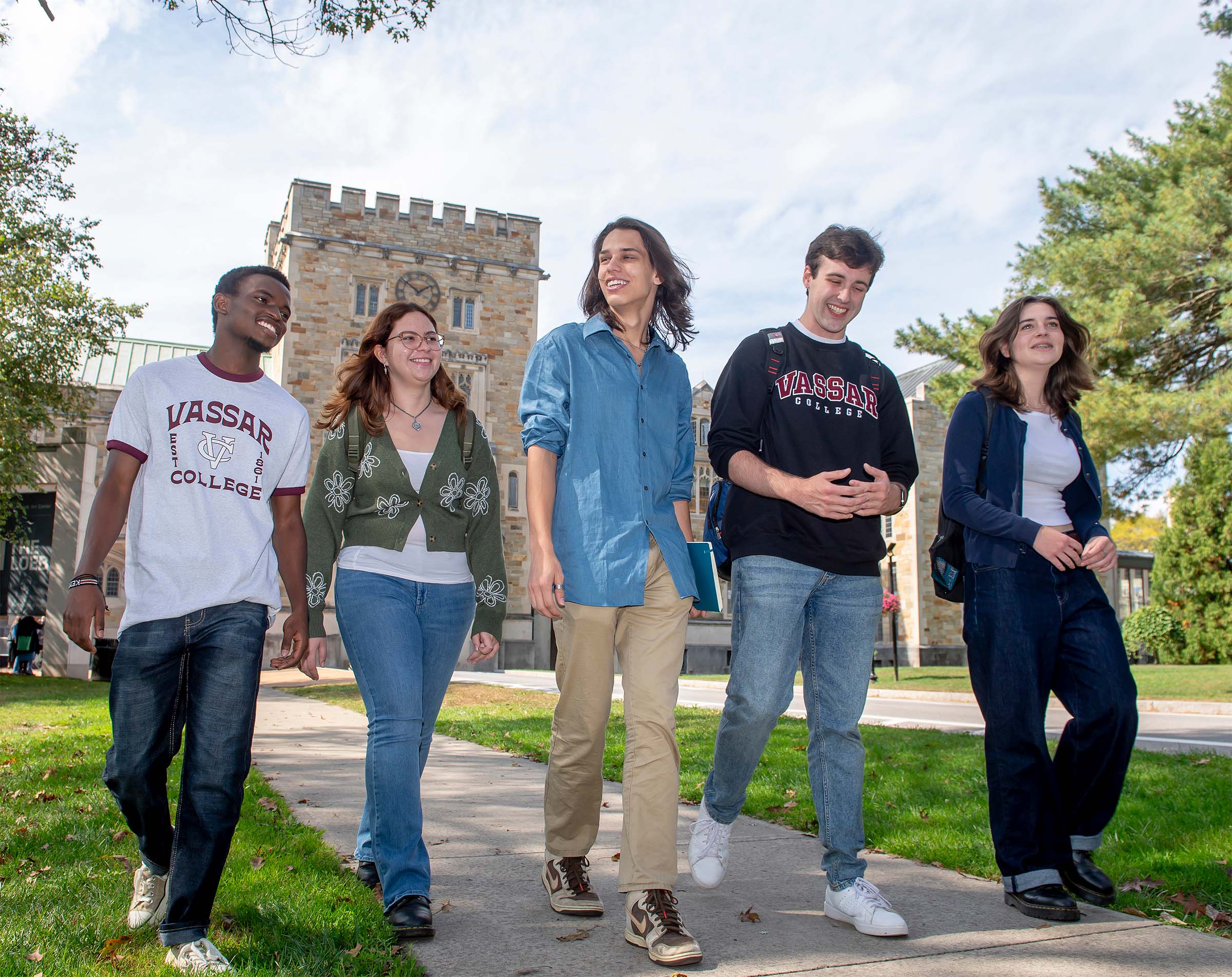 Five individuals walk on a campus sidewalk in front of a gothic-style stone building with a clock tower.