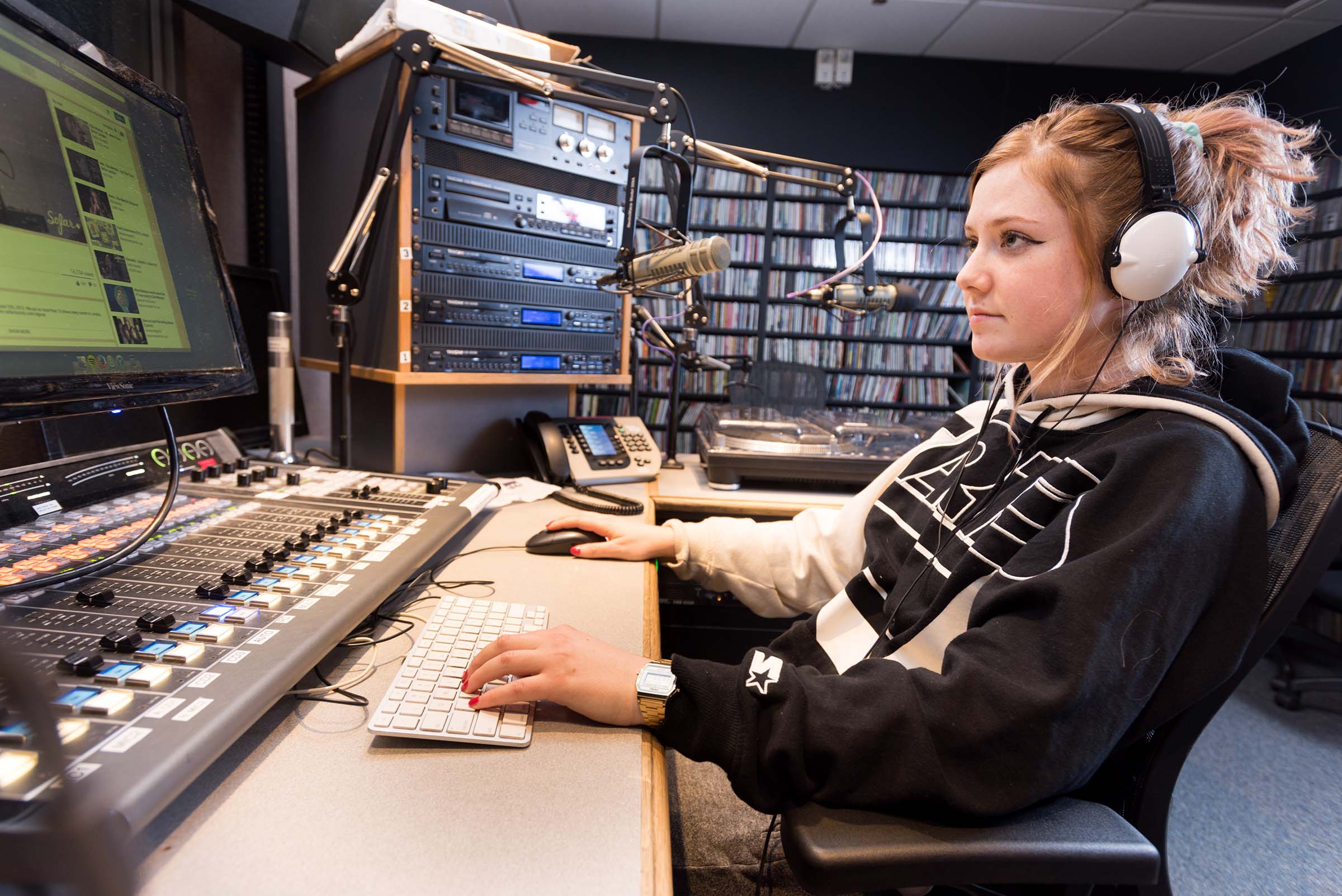 A person wearing headphones sits at a broadcasting console in a radio studio, using a computer keyboard and mouse in front of audio equipment and microphones.